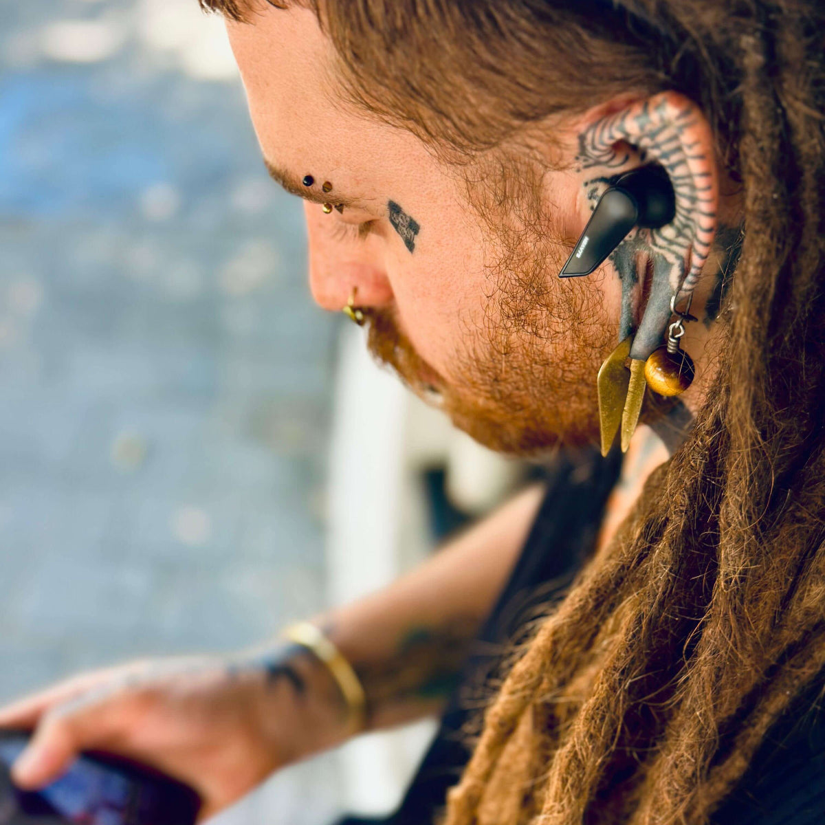 Person with dreadlocks and unique ear piercings using earbuds and phone outdoors